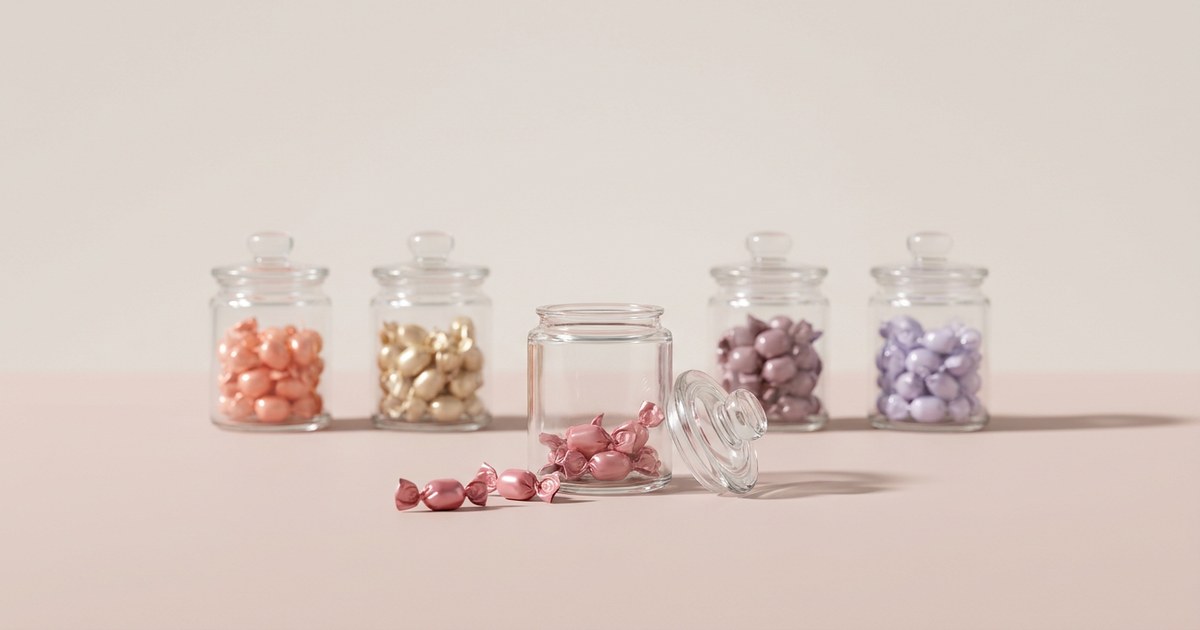Five glass apothecary jars in a row on a pale blush surface, the centre one open with its lid removed and most of its rose-pink twist-wrapped candies spilled out in front, while the four other jars stand closed and full of coral, champagne-gold, mauve, and lavender candies.