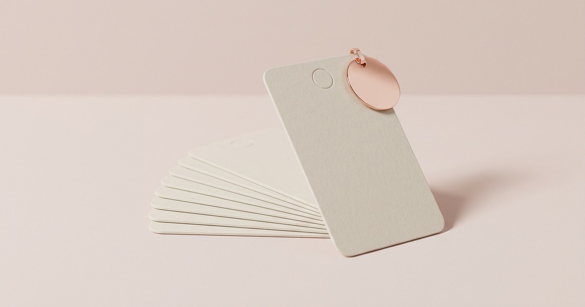 A fanned stack of eight identical blank cream cards with rounded corners lying on a pale blush surface, each with a small empty circle near the top, and the front card standing upright with a single polished rose gold disc tag clipped to its top corner.