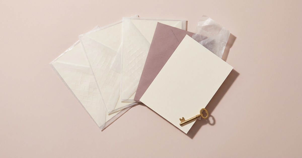 Overhead flat lay of blank cream cards fanned on a pale pink surface, three sealed in translucent sleeves with an embossed diamond lattice and one slid free, clean and unmarked, with a small brass key resting on it
